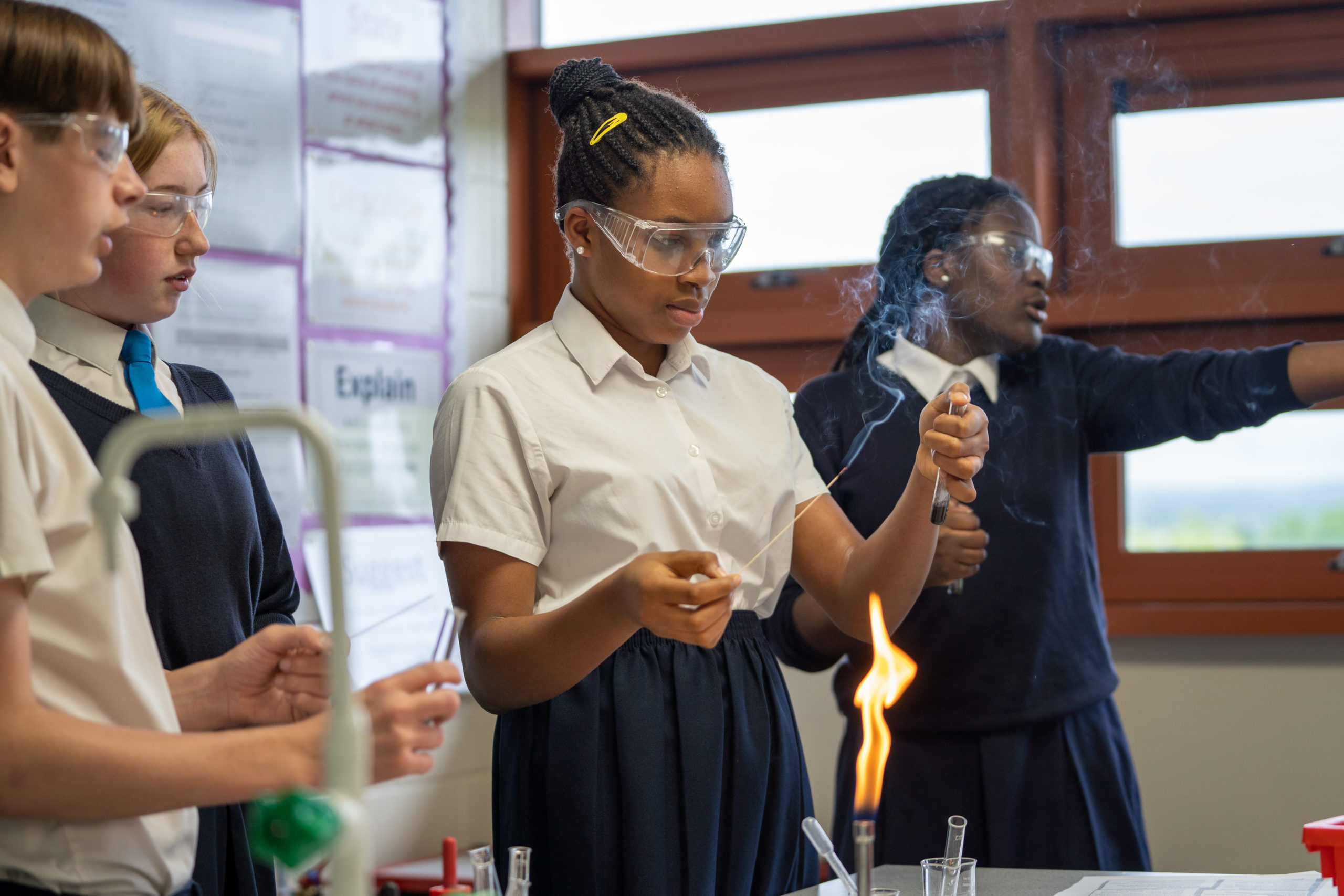 A small group of students are seen wearing safety goggles conducting an experiment involving Bunsen Burners in a Science Lab.