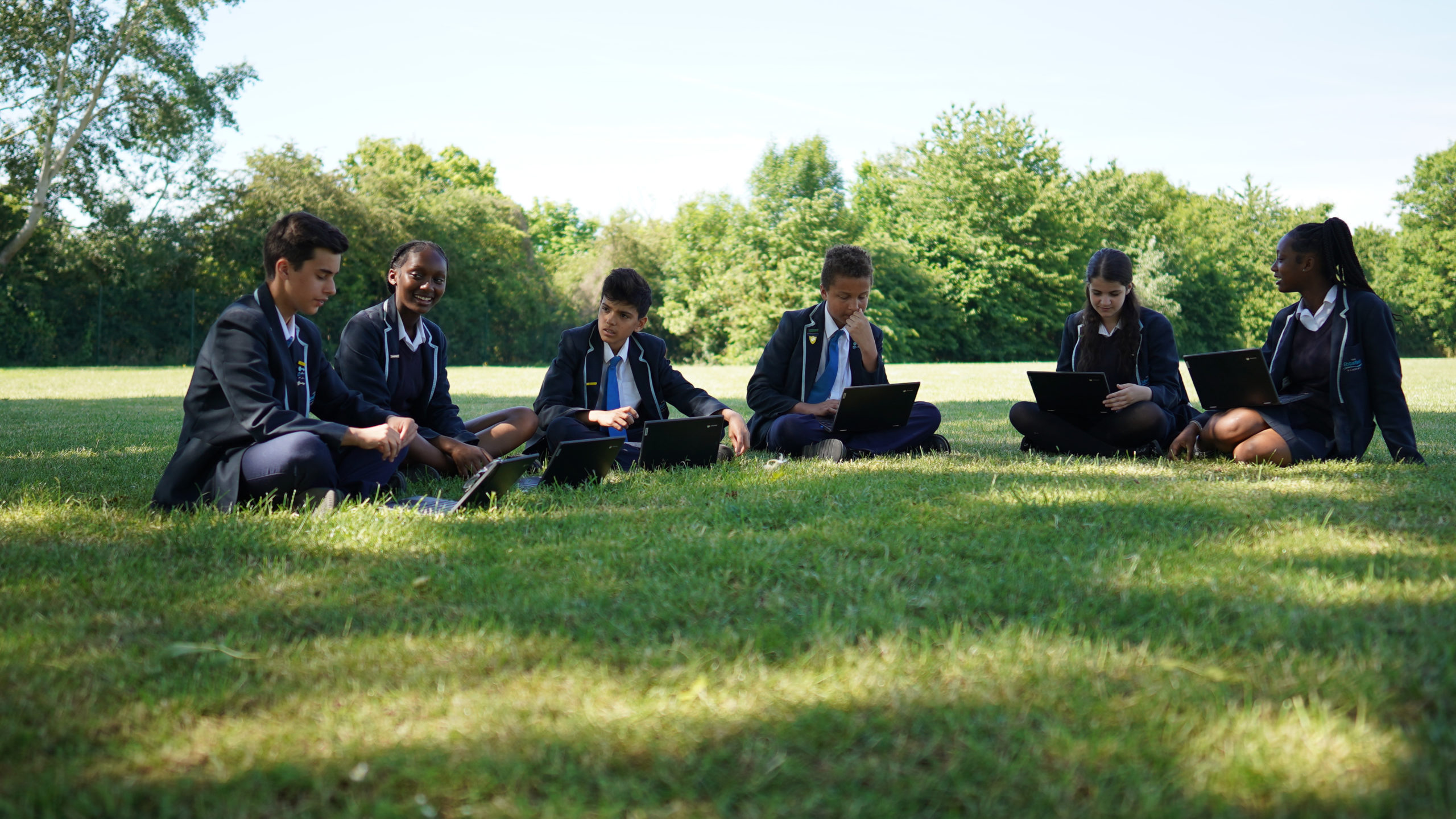 Six Ebbsfleet Academy students are seen sitting on a grass area outside and using their laptop computers.