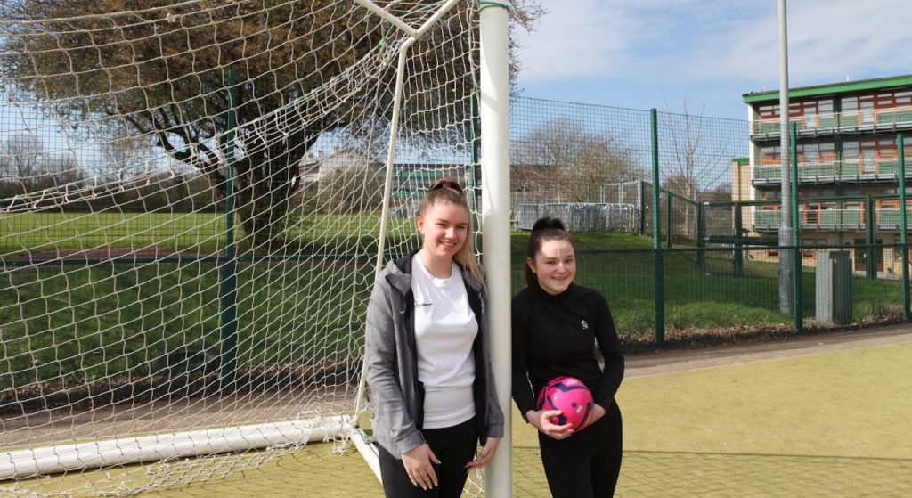 Two girls are shown posing for the camera beside a goalpost in celebration of Women's Football taking place at Ebbsfleet Academy.