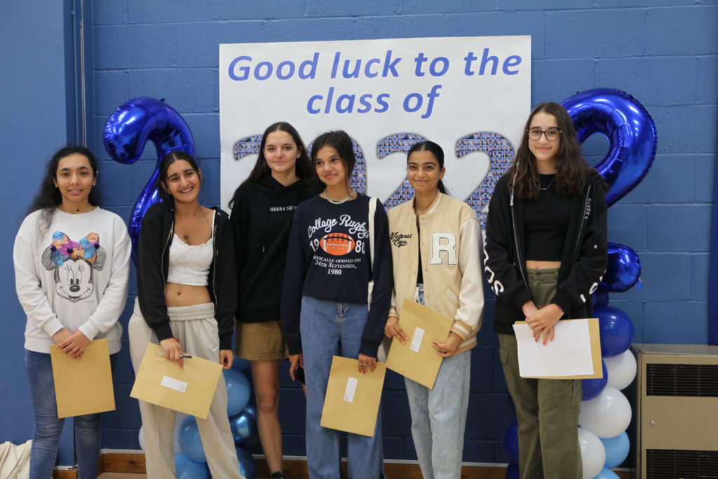 A small group of students are seen smiling for the camera, whilst holding their results envelopes on GCSE Results Day 2022.