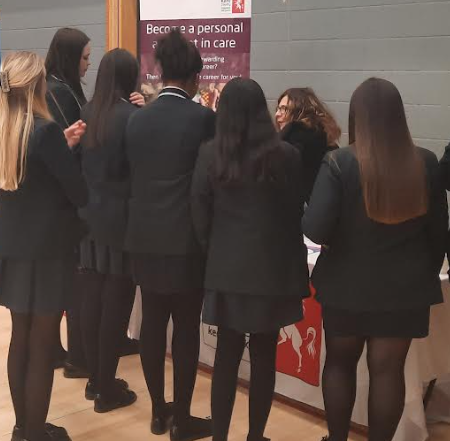 Photo showing a small group of academy students listening to a speaker behind a stall at an event in the Sports Hall.