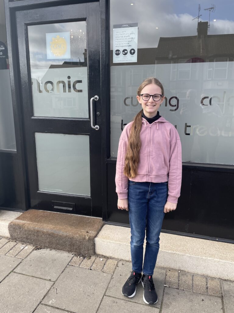 A female student is pictured standing beside the door to a hairdresser's salon with a smile on her face. She is seen wearing a pink hoodie and jeans.