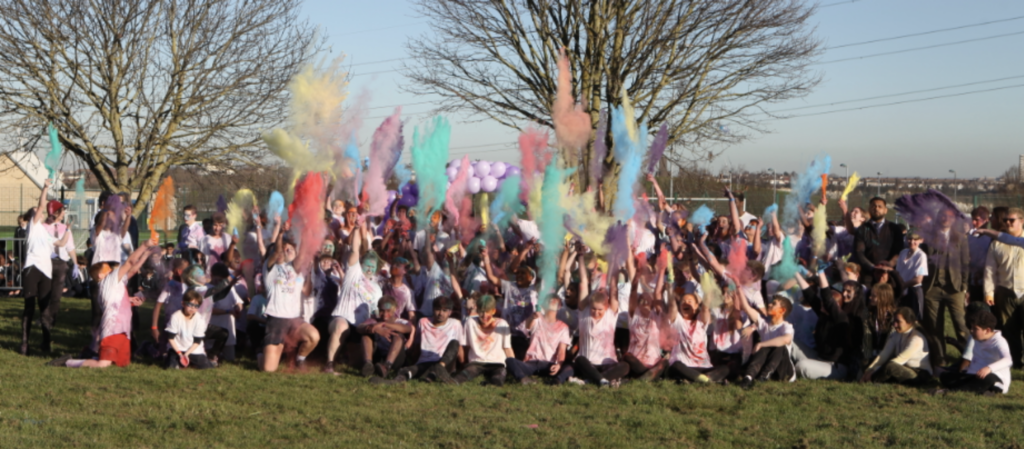 A large group of students are seen gathered together on the academy grounds, throwing multi-coloured powder into the air in celebration of the Hindu festival Holi.