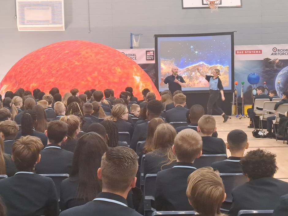 Students are seen gathered in the main hall, listening to a speaker from the company BAE Systems at the front of the room.