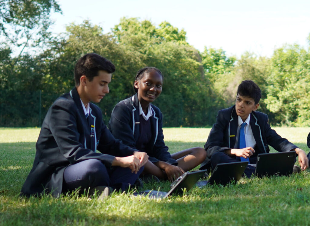 Three Ebbsfleet Academy students are pictured sitting together outdoors on the grass, open laptops in front of them and working together.