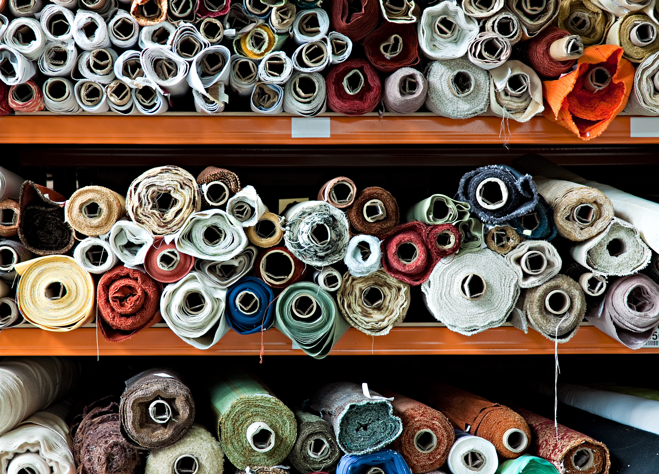 A large cabinet is seen with three shelves stacked full of different coloured rolls of textile materials.