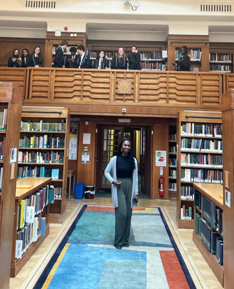 Year 11 and 12 students are pictured standing on a balcony area in the Library at Oxford University during a school trip there. A young female University student is seen stood, smiling at the camera in the area below the balcony.