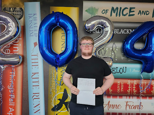 A male Post-16 student is seen smiling for the camera, whilst holding up his A-Level results.