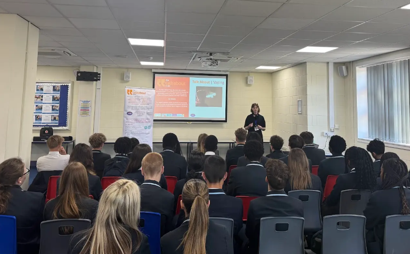 Teacher: On the left side of the room, a woman with dark hair and glasses is standing in front of a whiteboard, gesturing with her hands as she speaks. She appears to be leading a lesson. She is wearing dark clothing and has an ID badge. Students: A group of students, mostly teenagers, are seated at desks arranged in rows, facing the front of the classroom. Many of them are looking towards the teacher and the front of the room. Some are wearing school uniforms (dark blazers).