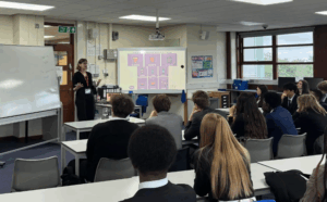 Here's a breakdown of what's in the picture: Teacher: On the left side of the room, a woman with dark hair and glasses is standing in front of a whiteboard, gesturing with her hands as she speaks. She appears to be leading a lesson. She is wearing dark clothing and has an ID badge. Students: A group of students, mostly teenagers, are seated at desks arranged in rows, facing the front of the classroom. Many of them are looking towards the teacher and the front of the room. Some are wearing school uniforms (dark blazers). Classroom Environment: Whiteboards: There's a large whiteboard on the left, partially filled with writing, and another large interactive whiteboard or screen at the front of the room displaying a presentation with illustrations (possibly related to "units" or measurements). Projector: A projector is mounted on the ceiling, pointed towards the main screen. Desks and Chairs: Individual desks and chairs are arranged for the students. Windows: Windows are visible on the right side of the room, letting in natural light. Posters/Decorations: Various posters and charts are on the walls, typical of a classroom. Door: A door with an "Exit" sign above it is visible in the background on the left. Supplies: Some school supplies or boxes are visible near the windows.