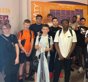 This picture shows a group of young people, likely boys, standing indoors. They appear to be at a trampoline park or similar recreation center, as suggested by the "GRAVITY TIMETABLE" sign in the background. Most of the individuals are looking towards the camera. They are dressed in casual clothing, including t-shirts, shorts, and trainers. Some are wearing backpacks. The "GRAVITY TIMETABLE" sign behind them displays a schedule with various sessions like "OPEN JUMP," "SEND SESSION," and "PARENT & TODDLER" for different days of the week.