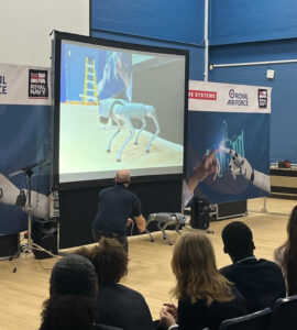 This picture shows an indoor event, likely a demonstration or presentation, taking place in what appears to be a hall or gymnasium with a wooden floor and blue walls. Here's a breakdown of what's happening: Main Focus: A man is on stage, interacting with a small, quadrupedal robot that resembles a dog. He is holding a leash or cable connected to the robot. Projector Screen: Behind the man, a large screen displays a close-up video feed of a similar or the same type of robot in action. Audience: Several individuals, including some with their backs to the camera, are seated on chairs in front of the stage, watching the demonstration. Banners: In the background, there are banners with "ROYAL NAVY" and "ROYAL AIR FORCE" logos, suggesting the event might be related to military technology, recruitment, or a STEM outreach program supported by these organizations. There's also a "BAE SYSTEMS" logo visible, indicating their involvement.