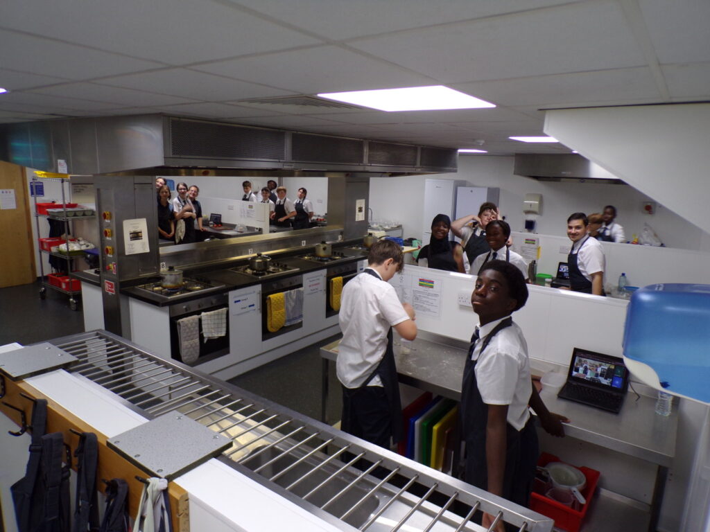 This photo provides a broad view of the kitchen space, showing multiple stations where students are working. Some students are at the counters preparing ingredients, while others appear to be standing by ovens or sinks. Several laptops are visible on the counters, suggesting they might be following recipes or instructions electronically. The overall atmosphere seems to be one of busy, collaborative learning.