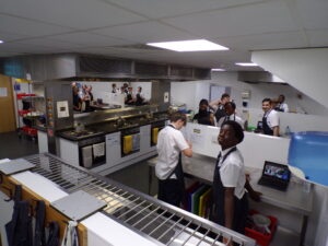 This photo provides a broad view of the kitchen space, showing multiple stations where students are working. Some students are at the counters preparing ingredients, while others appear to be standing by ovens or sinks. Several laptops are visible on the counters, suggesting they might be following recipes or instructions electronically. The overall atmosphere seems to be one of busy, collaborative learning.