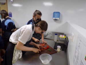 Two female students are working together at a counter, both actively chopping tomatoes on a cutting board. They are wearing aprons, and there are various kitchen supplies around them