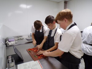 Three male students are closely gathered around a cutting board, collaborating on chopping tomatoes and other vegetables. They are all wearing aprons and focused on the task at hand.