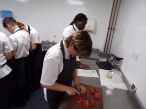 A female student in the foreground is focused on chopping tomatoes on a cutting board. Other students are visible in the background, also engaged in kitchen tasks. A laptop is open on the counter next to the student, possibly displaying a recipe.