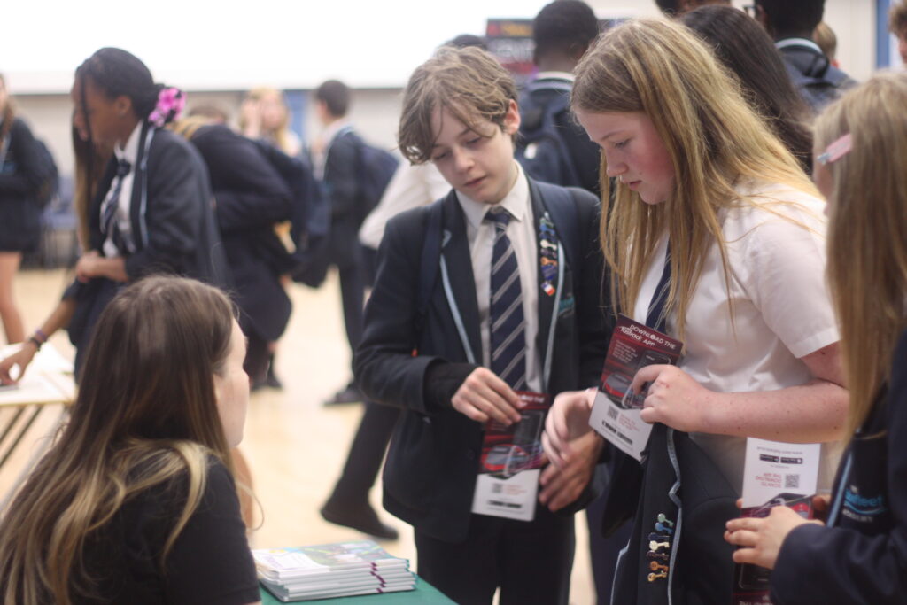 Several students in school uniforms are gathered around a table. Two students in the foreground are looking at some leaflets or papers, while another person sits at the table, seemingly assisting them. Other students are visible in the background, suggesting an event or fair.