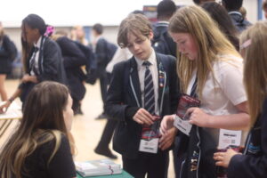 Several students in school uniforms are gathered around a table. Two students in the foreground are looking at some leaflets or papers, while another person sits at the table, seemingly assisting them. Other students are visible in the background, suggesting an event or fair.